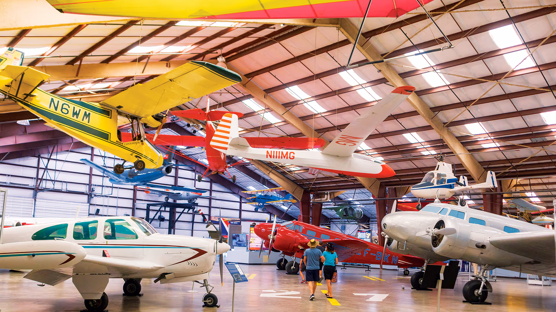 Interior view of an aviation museum hangar with various vintage airplanes displayed and two people walking down the center aisle.